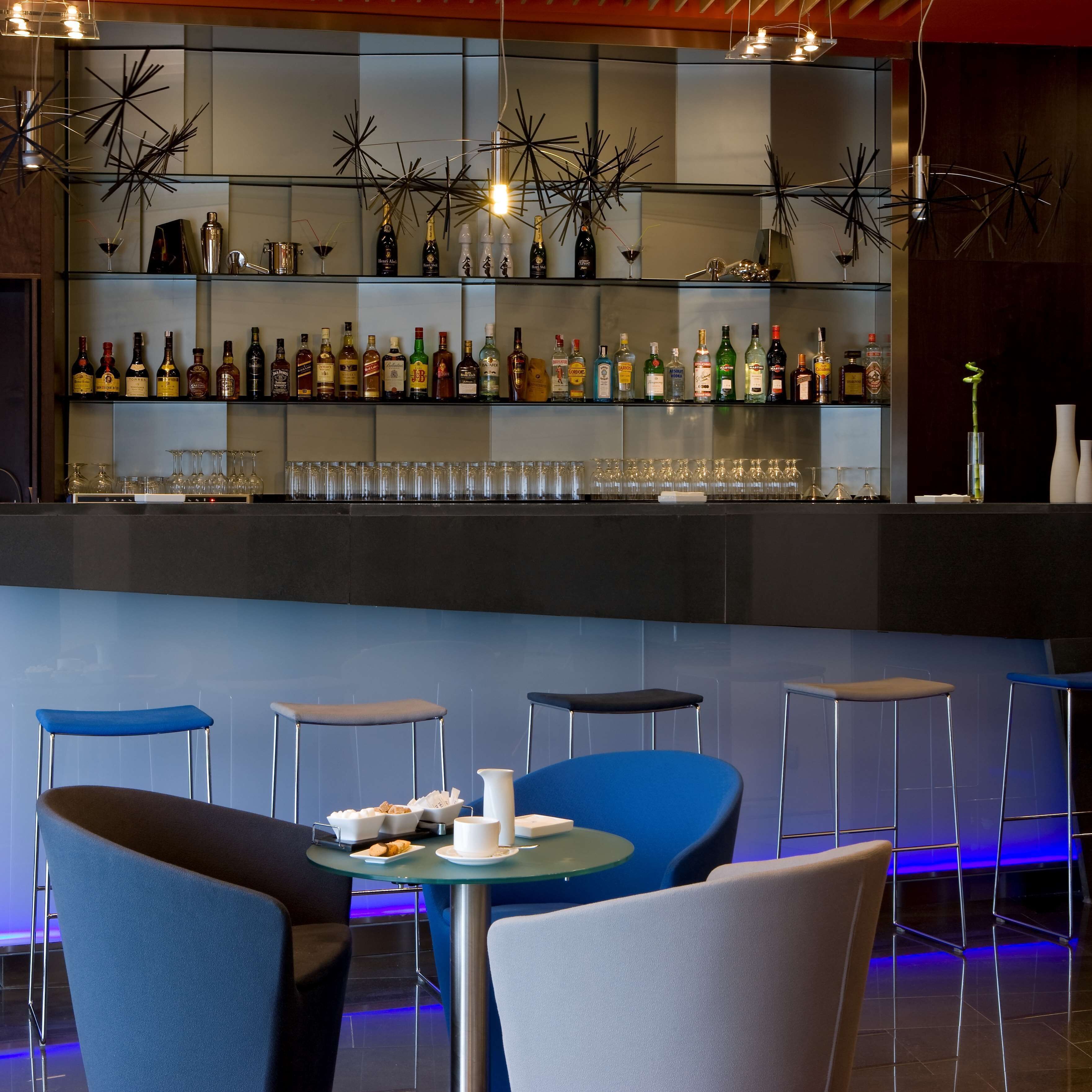Zona de bar moderna con una variedad de botellas de licores y vasos, taburetes azules y un área de estar con mesa y sillas.