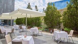 Un área de comedor al aire libre con mesas y sillas, un paraguas grande y un edificio al fondo.