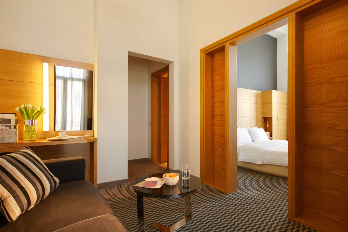 Habitación de hotel con sofá, una mesita con frutero y vasos, y una cama al fondo.