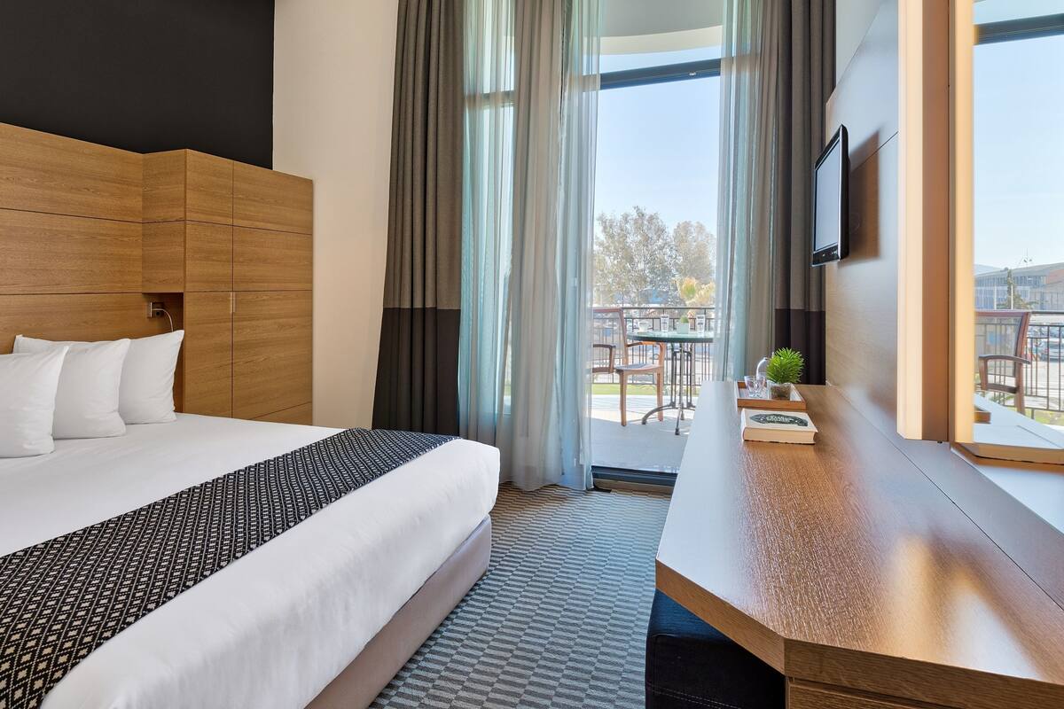 Habitación de hotel con una cama grande, cabecero de madera, escritorio con planta y vistas al exterior.