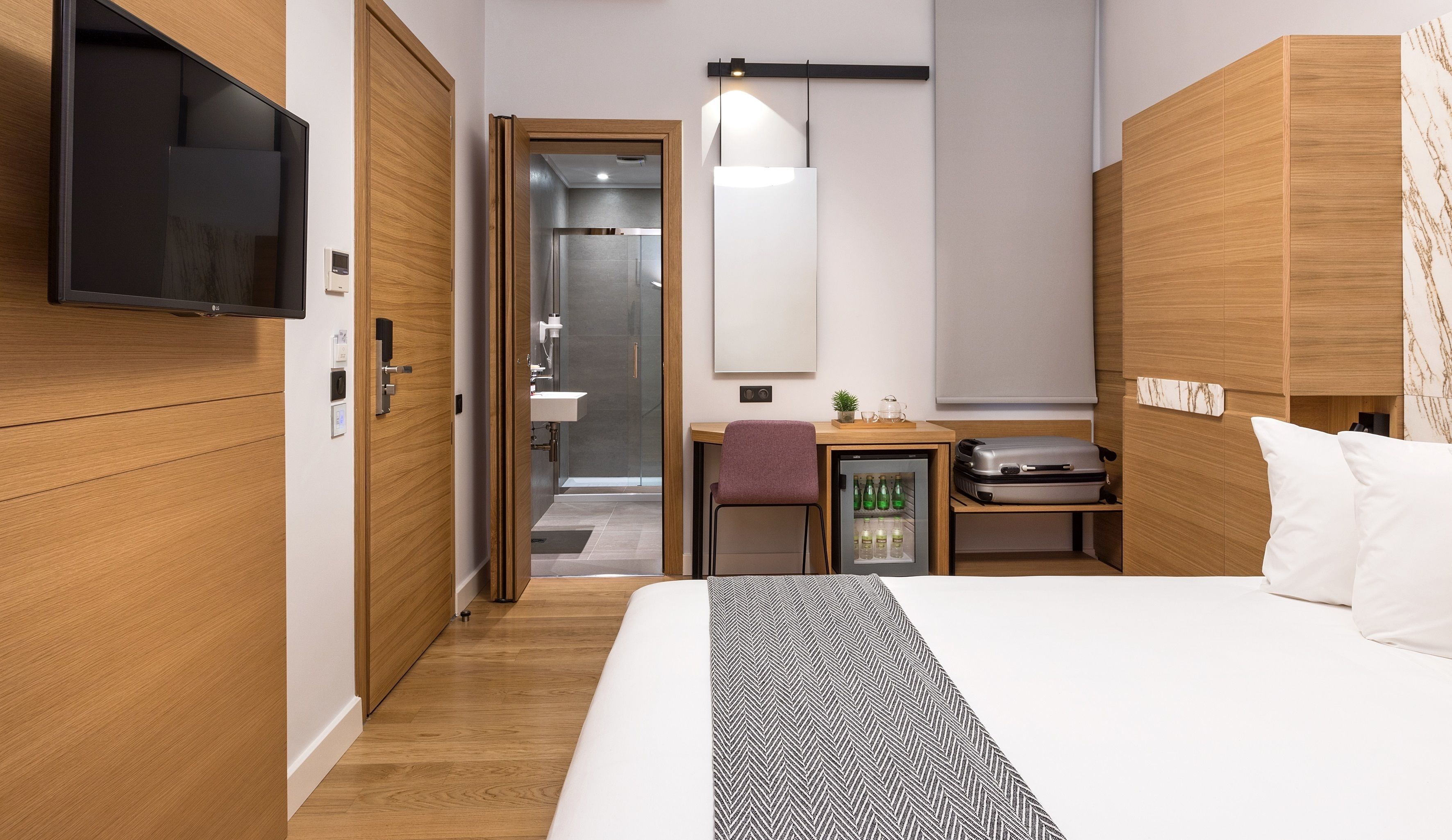 A hotel room with a bed, a desk with a chair, a television, a minibar, and a wardrobe.