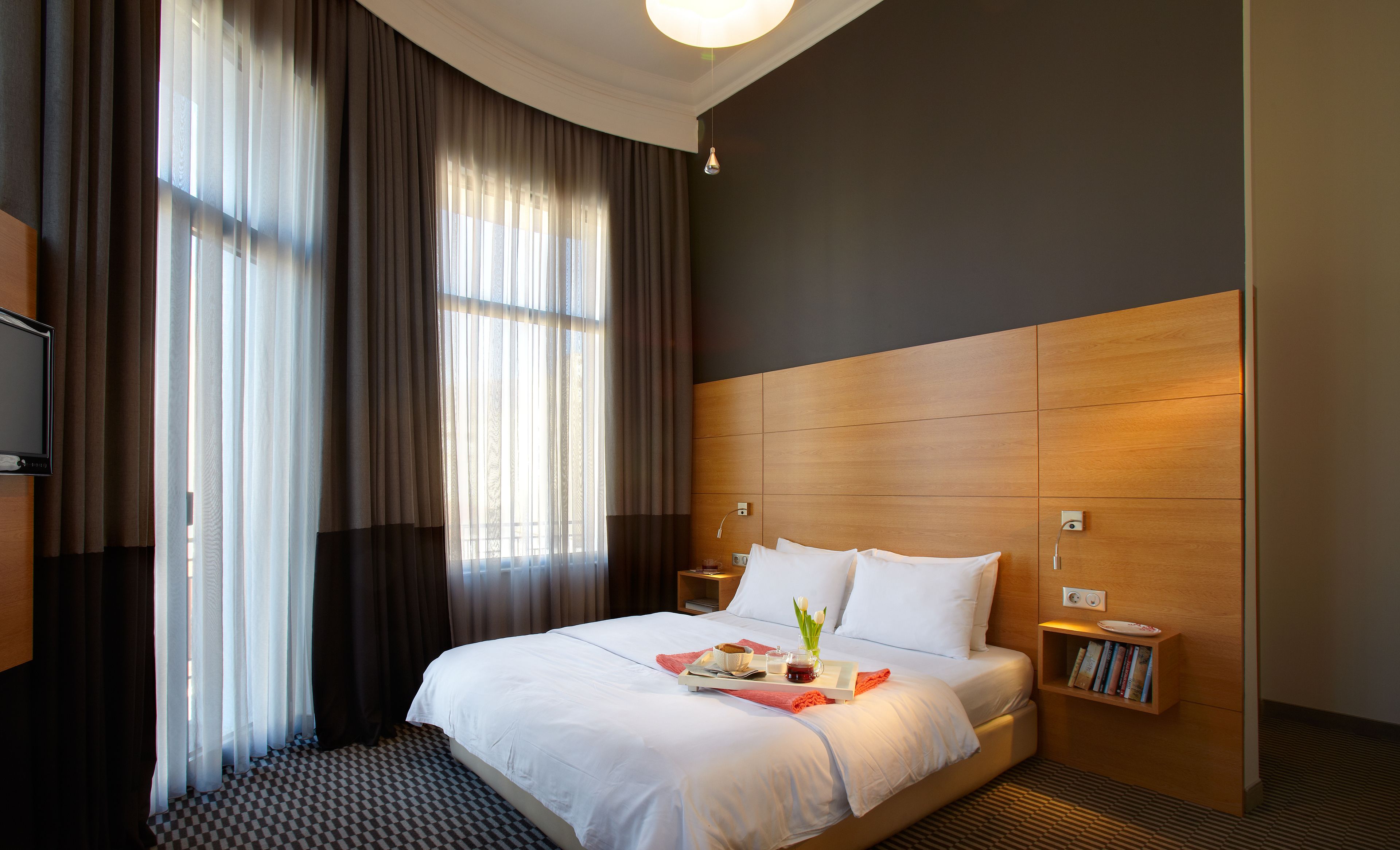 A hotel room with a large bed, a bedside table, a nightstand, a TV, and a window with curtains.