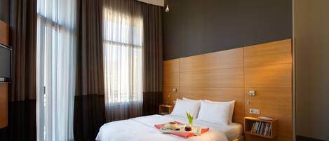 A hotel room with a large bed, a bedside table, a nightstand, a TV, and a window with curtains.