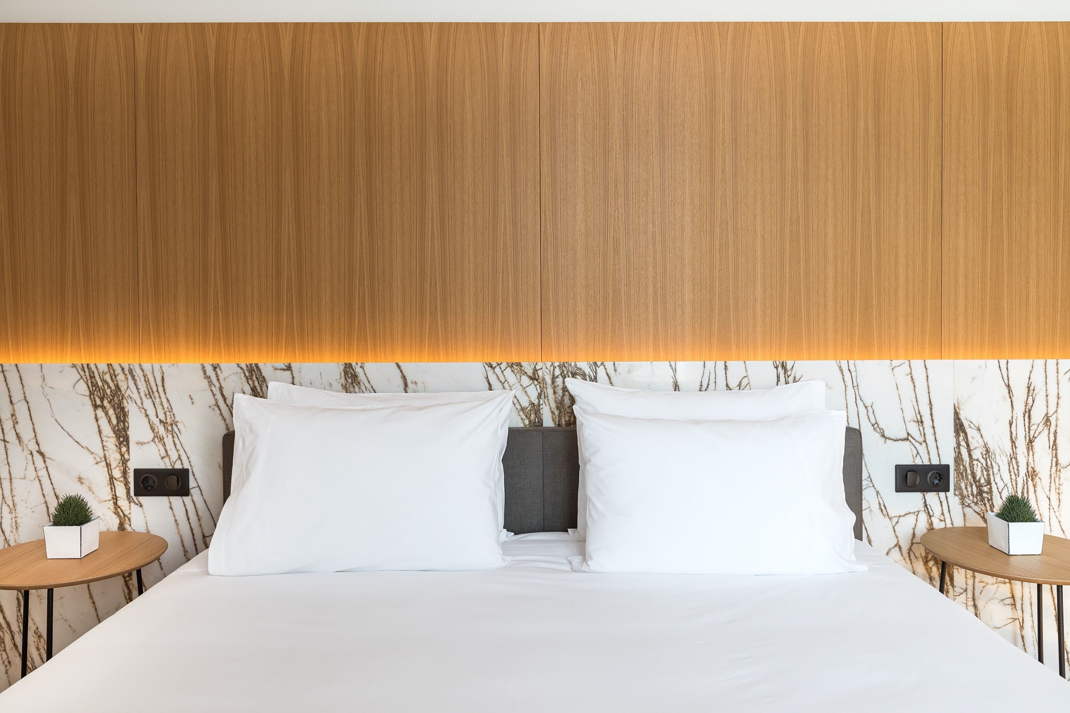 A modern hotel room with a large bed, marble-patterned walls, and a wooden headboard. There are two bedside tables with small potted plants.