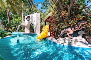 A pool area with a dinosaur statue, a waterfall feature, and lush tropical vegetation.