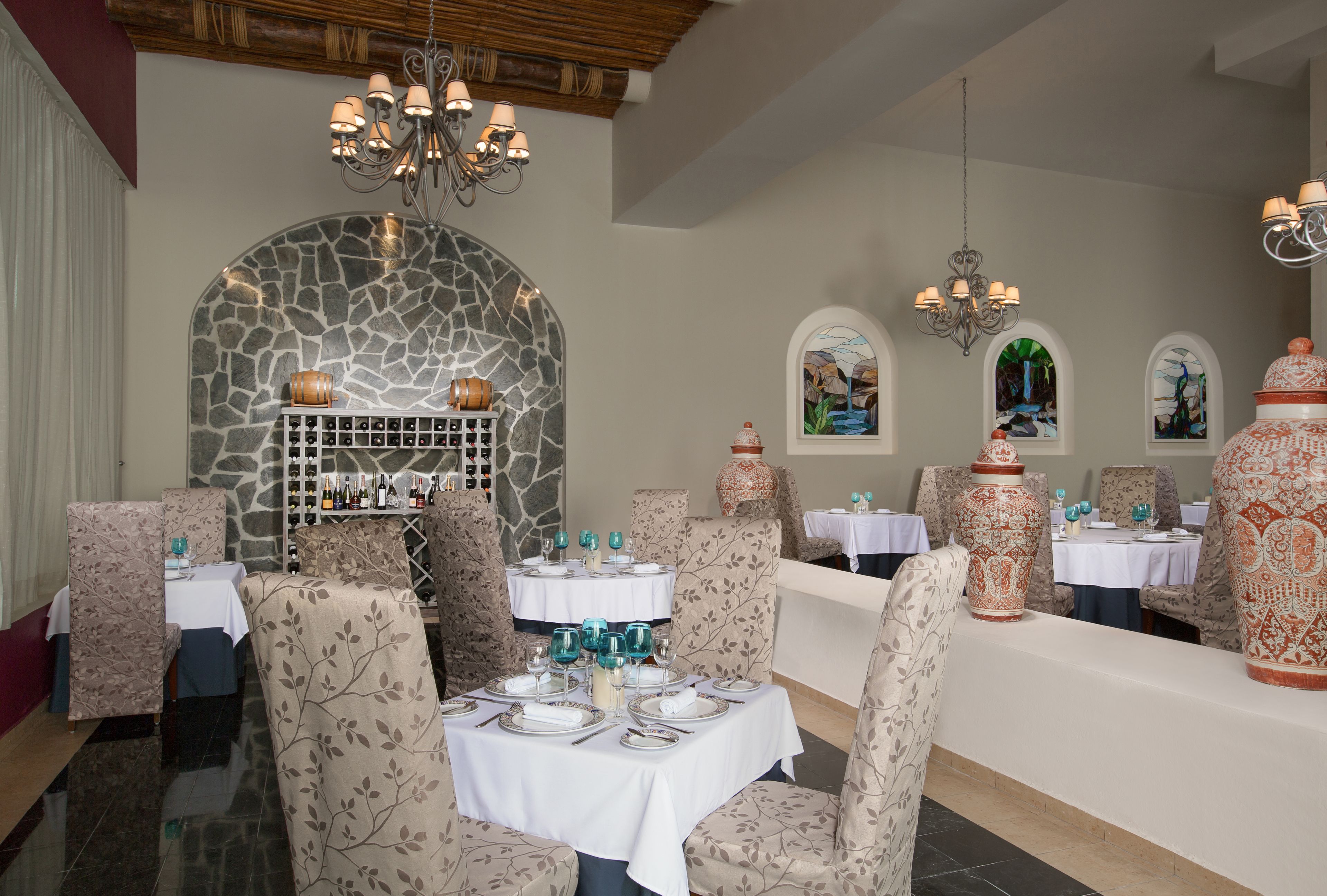 Restaurante com parede de pedra, lustres e cadeiras estampadas. Mesas montadas com toalhas brancas e talheres.