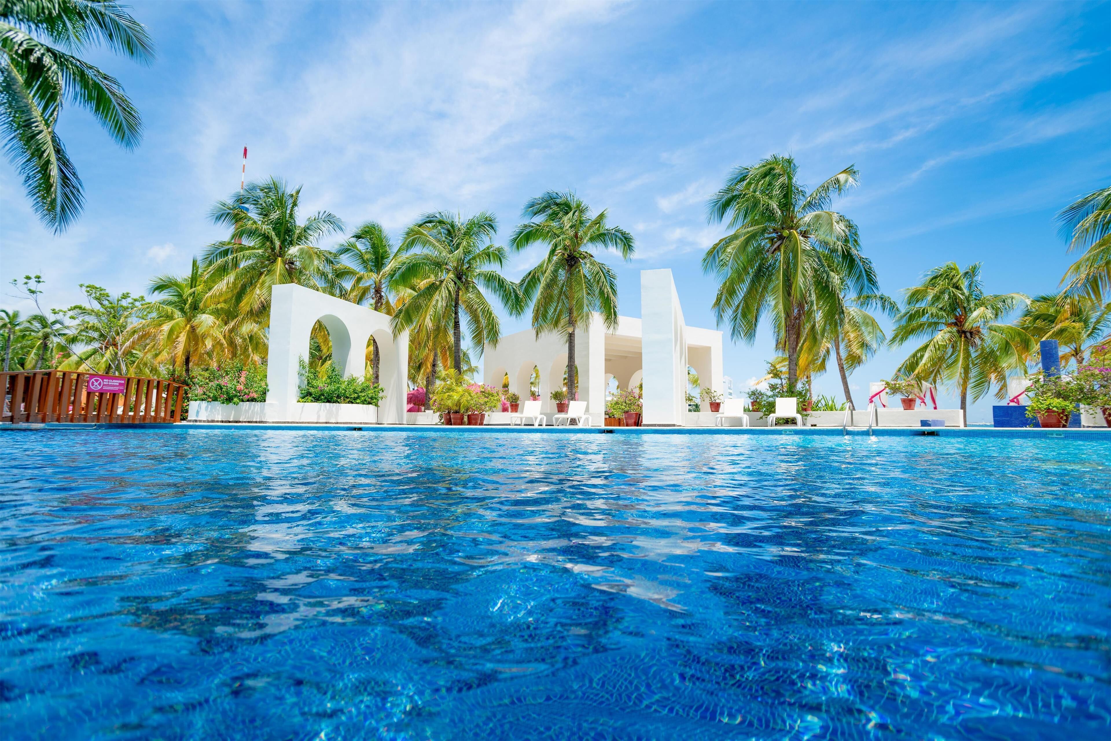 Una piscina con agua azul cristalina, rodeada de palmeras y un edificio blanco con arcos.