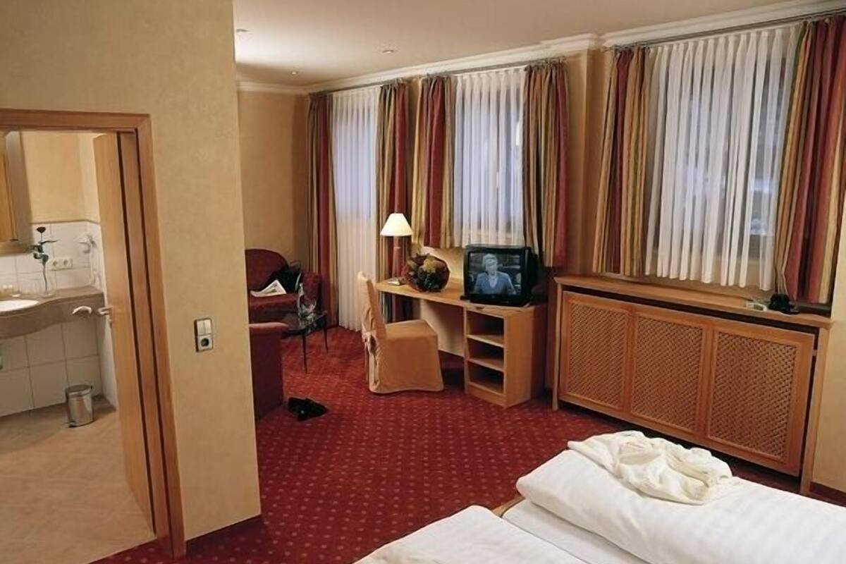 A hotel room with a bed, a desk with a TV, a chair, a small table with a lamp, a window with curtains, and a bathroom visible through an open door.