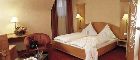 A hotel room with a large bed, a desk with a lamp, a chair, and a small table with glasses.