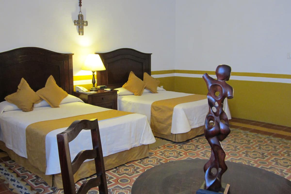 Habitación de hotel con dos camas, una mesita de noche, una lámpara, una silla de madera y una escultura decorativa.