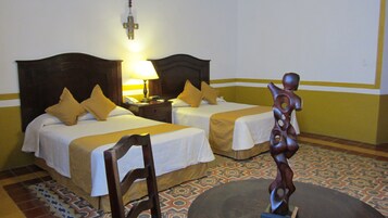 Habitación de hotel con dos camas, una mesita de noche, una lámpara, una silla de madera y una escultura decorativa.