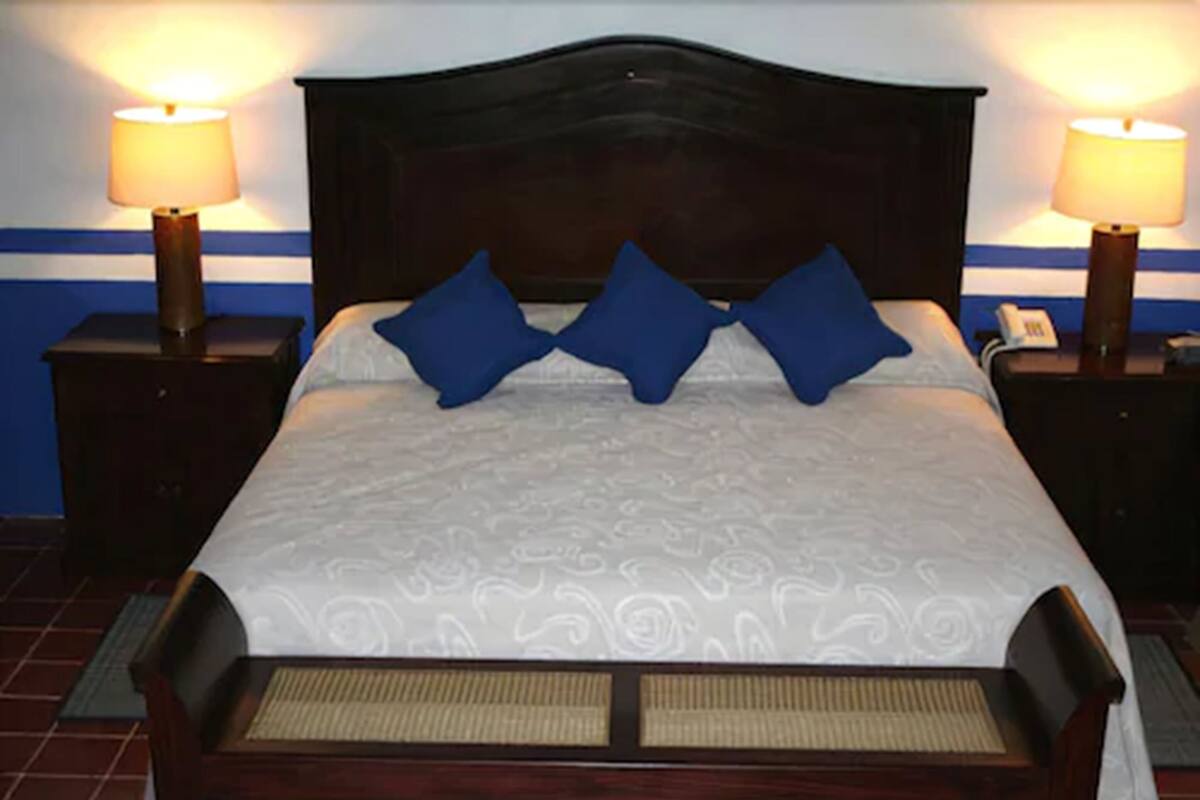 Una cama bien hecha con una colcha estampada y dos almohadas azules, flanqueada por dos mesitas de noche con lámparas, sobre un fondo de pared azul y blanca.