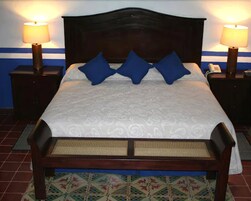 Una cama bien hecha con una colcha estampada y dos almohadas azules, flanqueada por dos mesitas de noche con lámparas, sobre un fondo de pared azul y blanca.