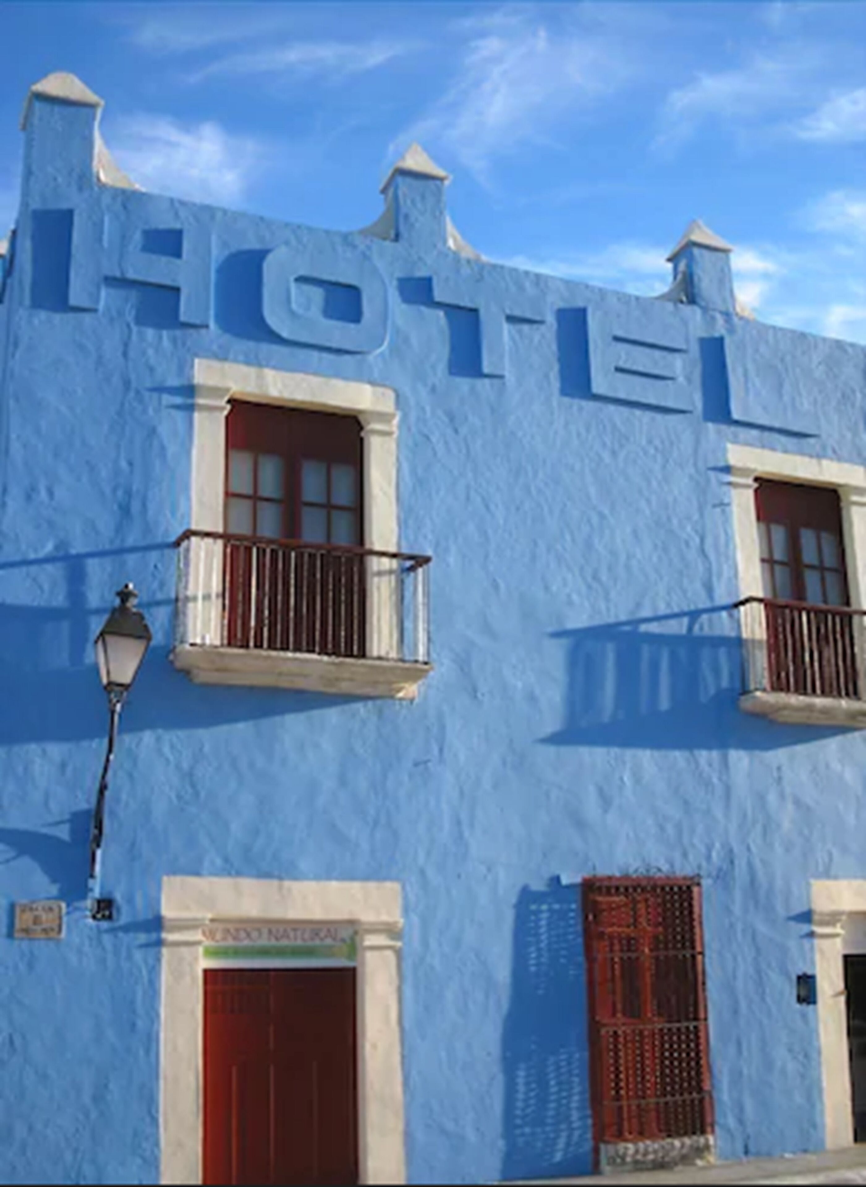 Een hotelgevel met een blauwe buitenzijde, twee ramen met balkons, een rode deur en een bord waarop 
