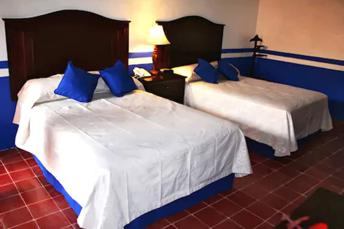 Habitación con dos camas, cada una con ropa blanca y almohadas azules, una mesita de noche con lámpara, y una lámpara fijada a la pared.