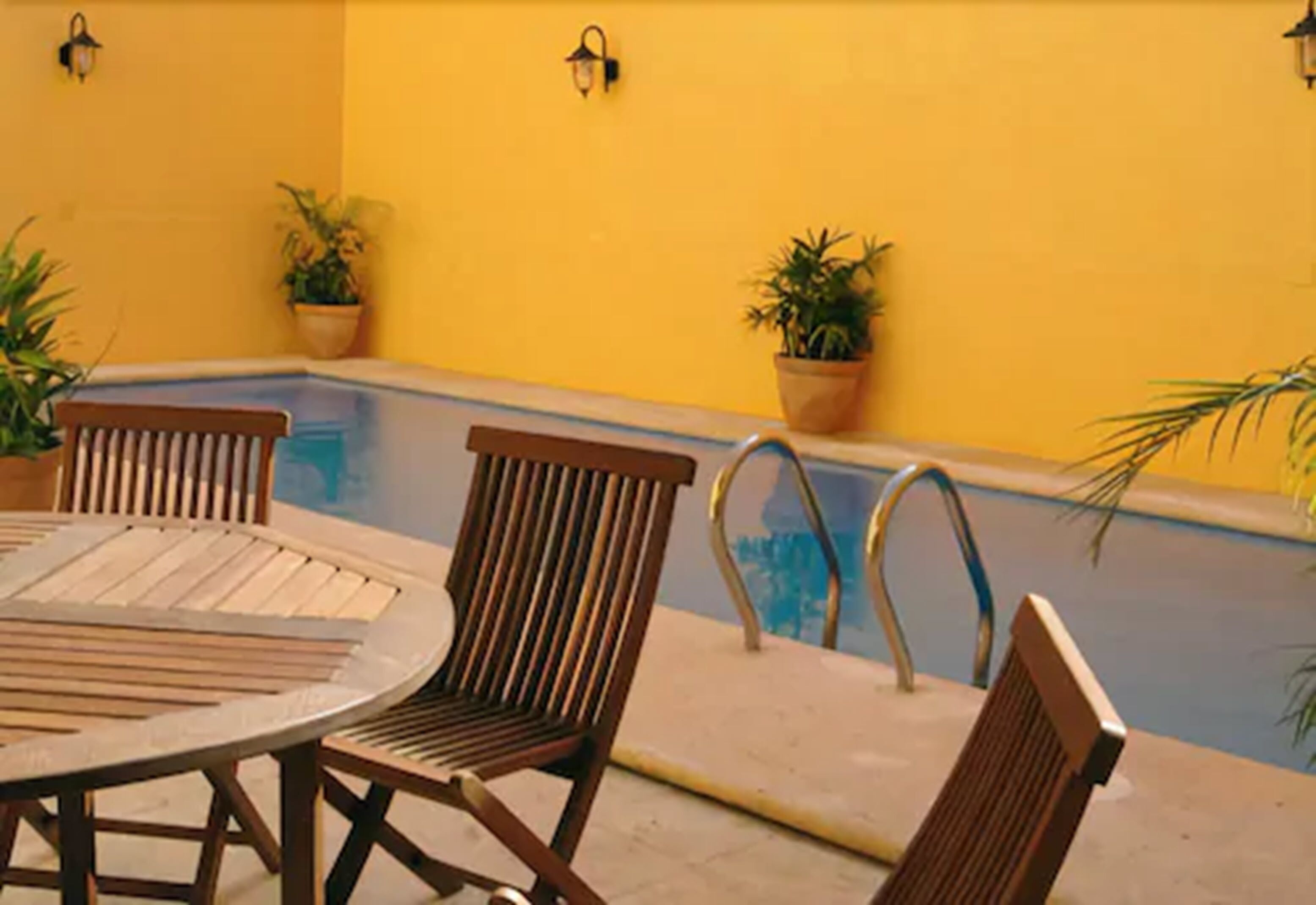 Zona junto a la piscina con una mesa redonda y cuatro sillas de madera, rodeada de paredes amarillas y plantas en macetas.
