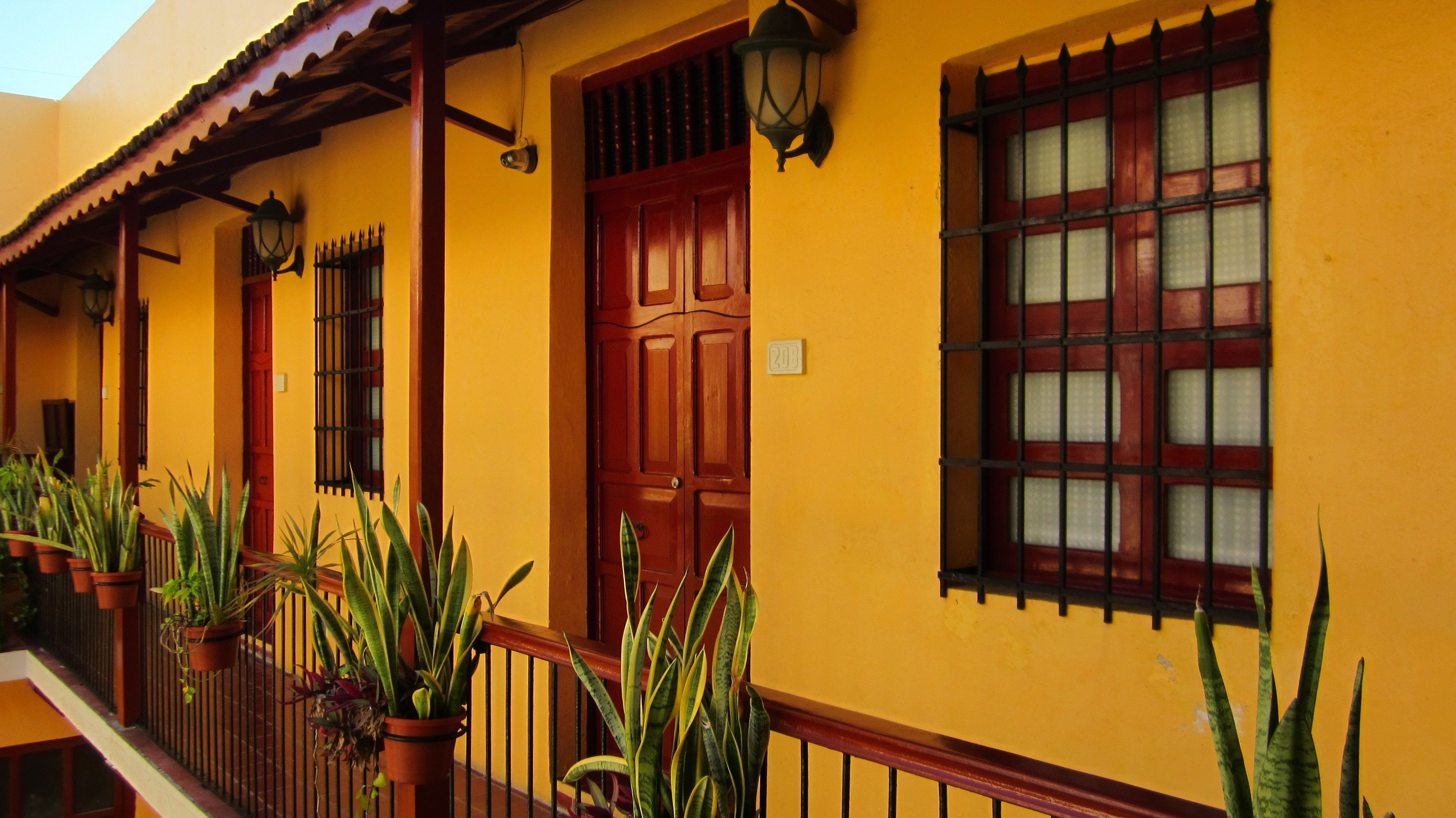 A yellow building with a wooden door, a window with bars, and potted plants along the railing.