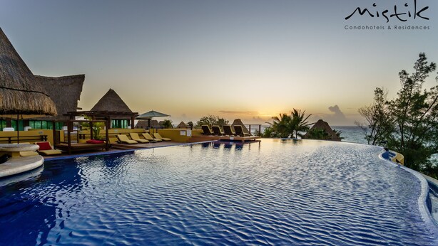 A poolside view with lounge chairs, a thatched-roof structure, and a sunset in the background. - Pueblito Escondido Luxury by Mistik (Playa del Carmen)