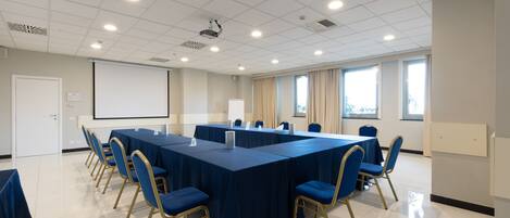 A conference room with a long table, blue chairs, a projector screen, and large windows.