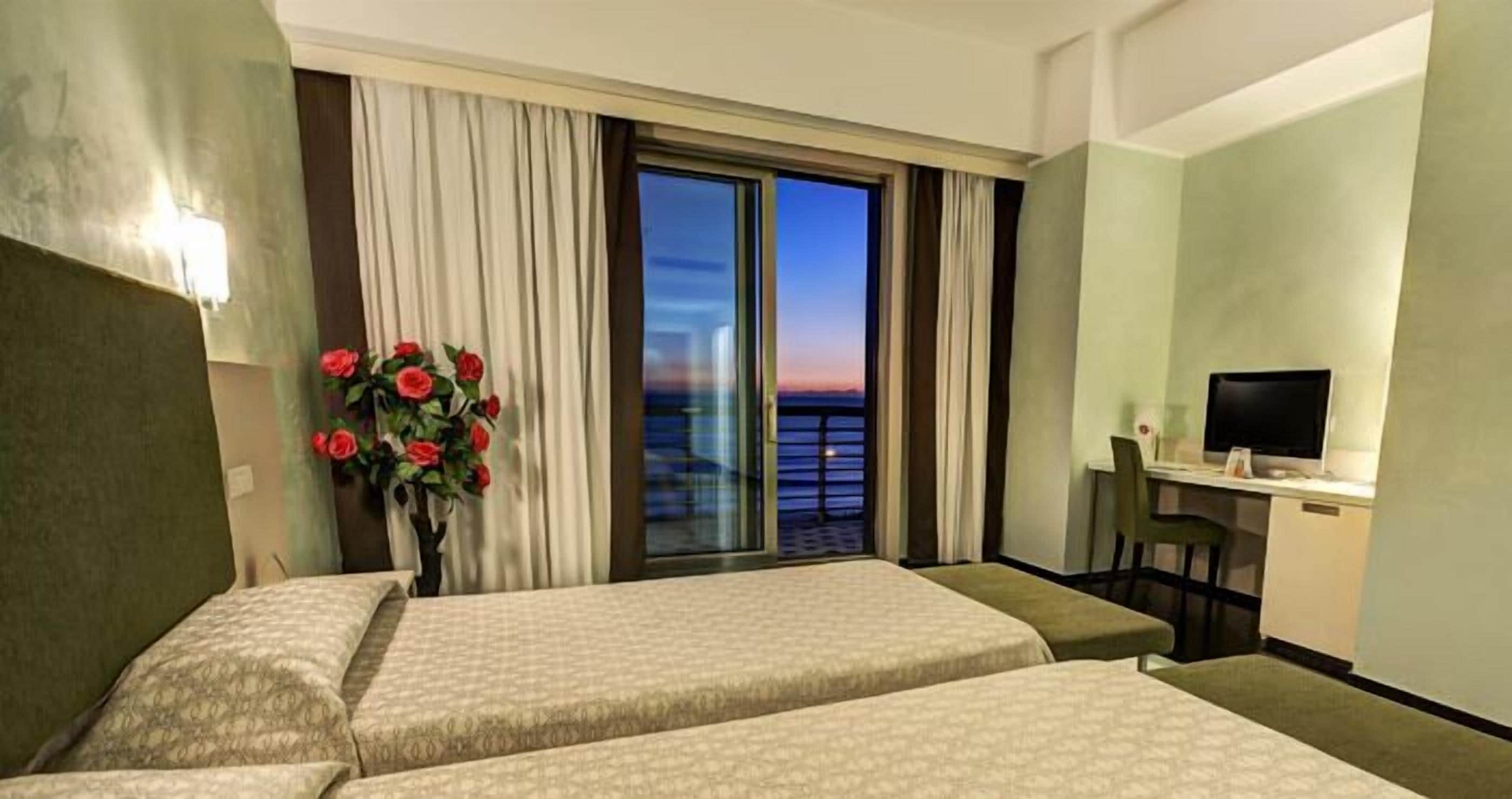 Habitación de hotel con dos camas, un escritorio con silla, televisión y vista al atardecer por la ventana.