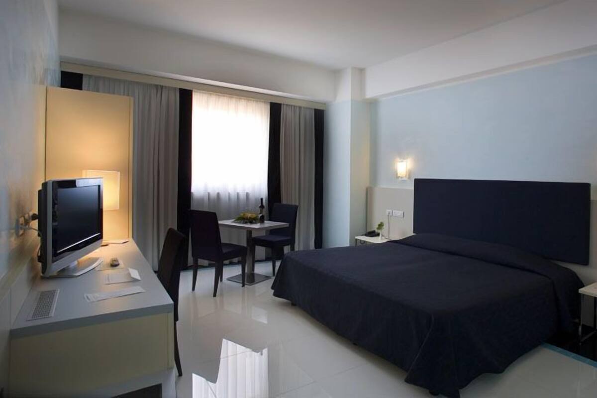 Una habitación de hotel con una cama, un escritorio con una computadora, una silla y una mesita con una lámpara.