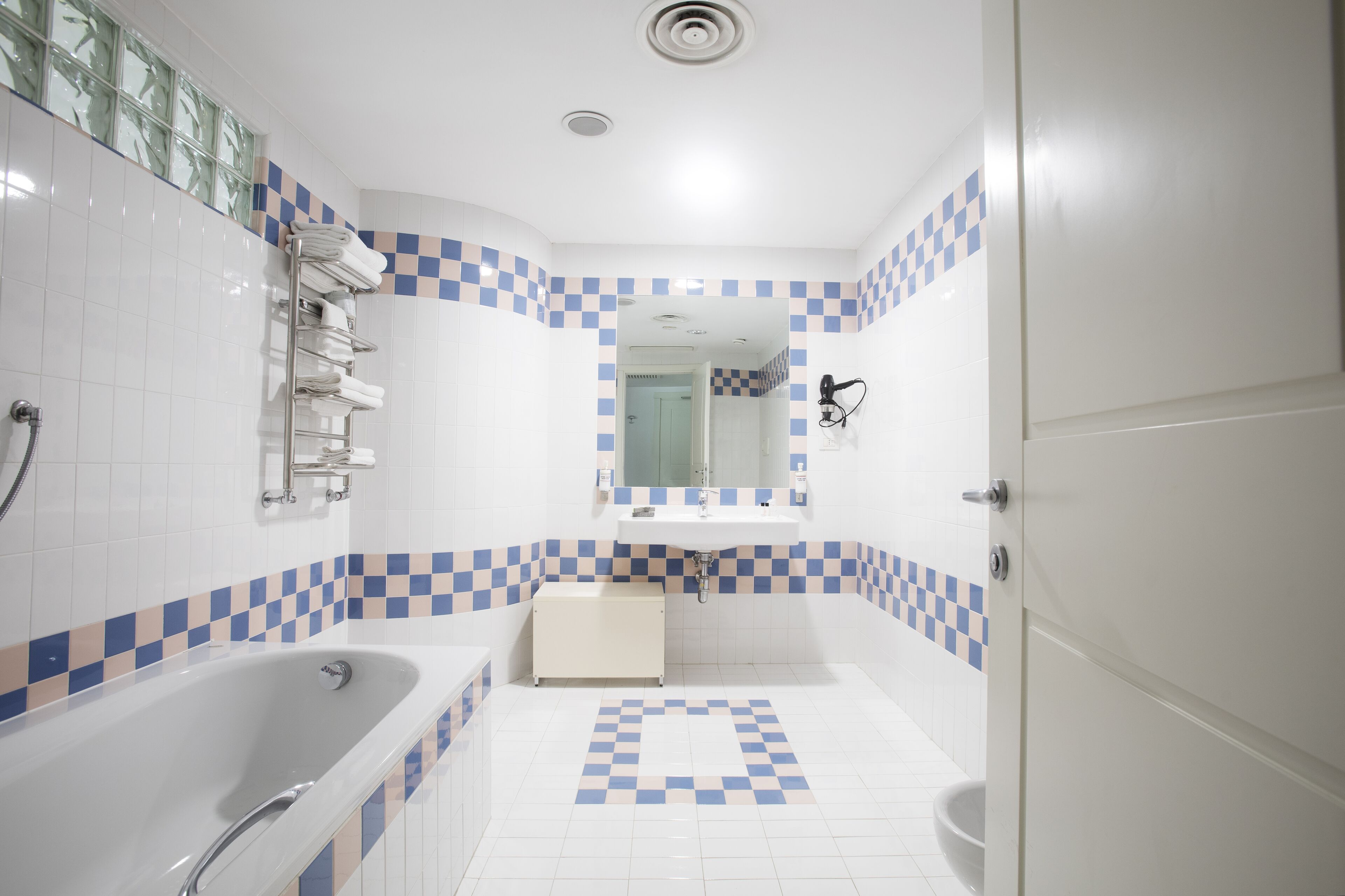 Un bagno con una vasca da bagno bianca, un lavandino e un water. È presente un portasciugamani con asciugamani piegati, uno specchio sopra il lavandino e un pavimento a scacchi.