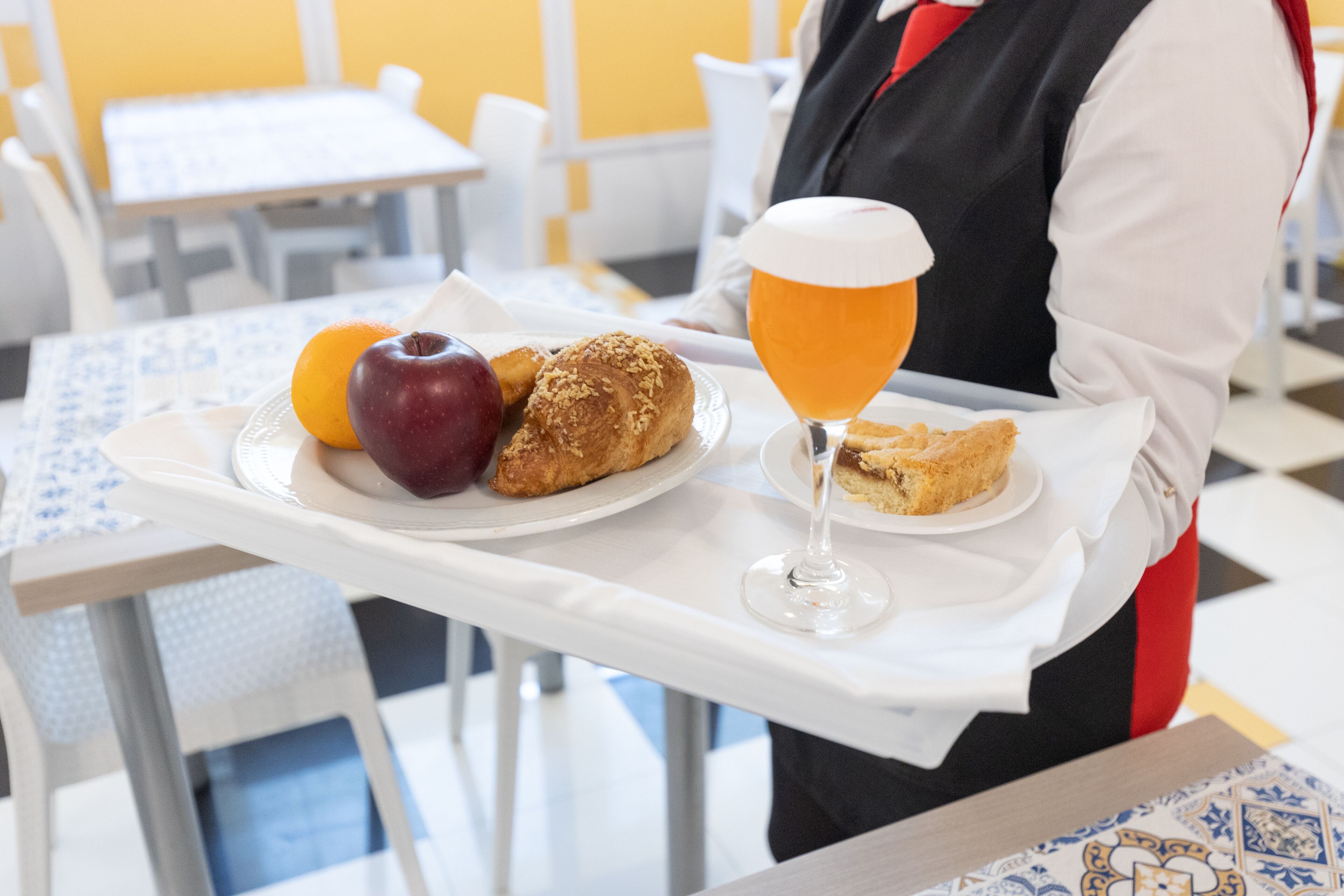 A tray with food items including an apple, a croissant, and a glass of beer, held by a person in a uniform.
