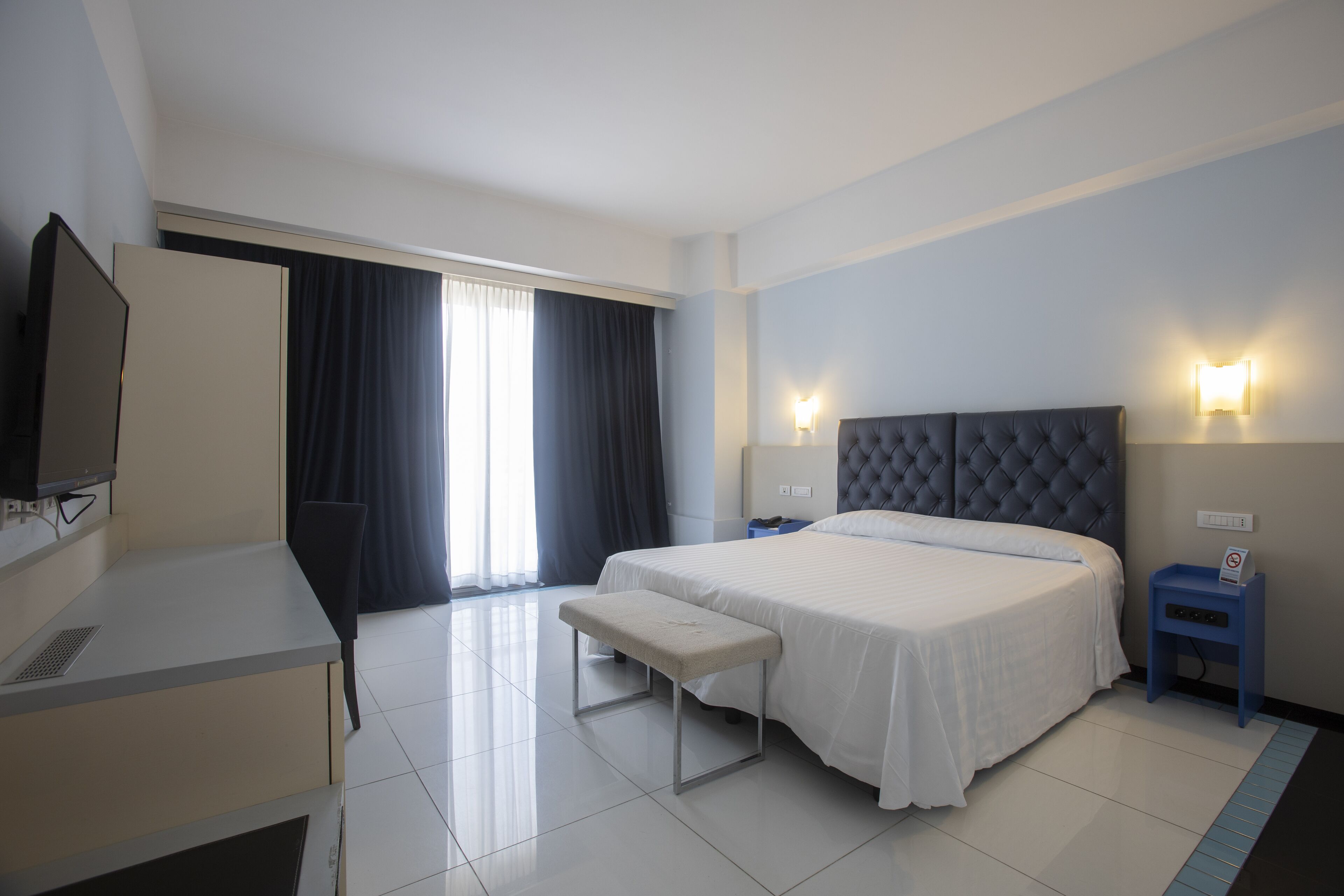 A hotel room with a bed, a desk, a chair, a bench, a TV, and a nightstand with a lamp.