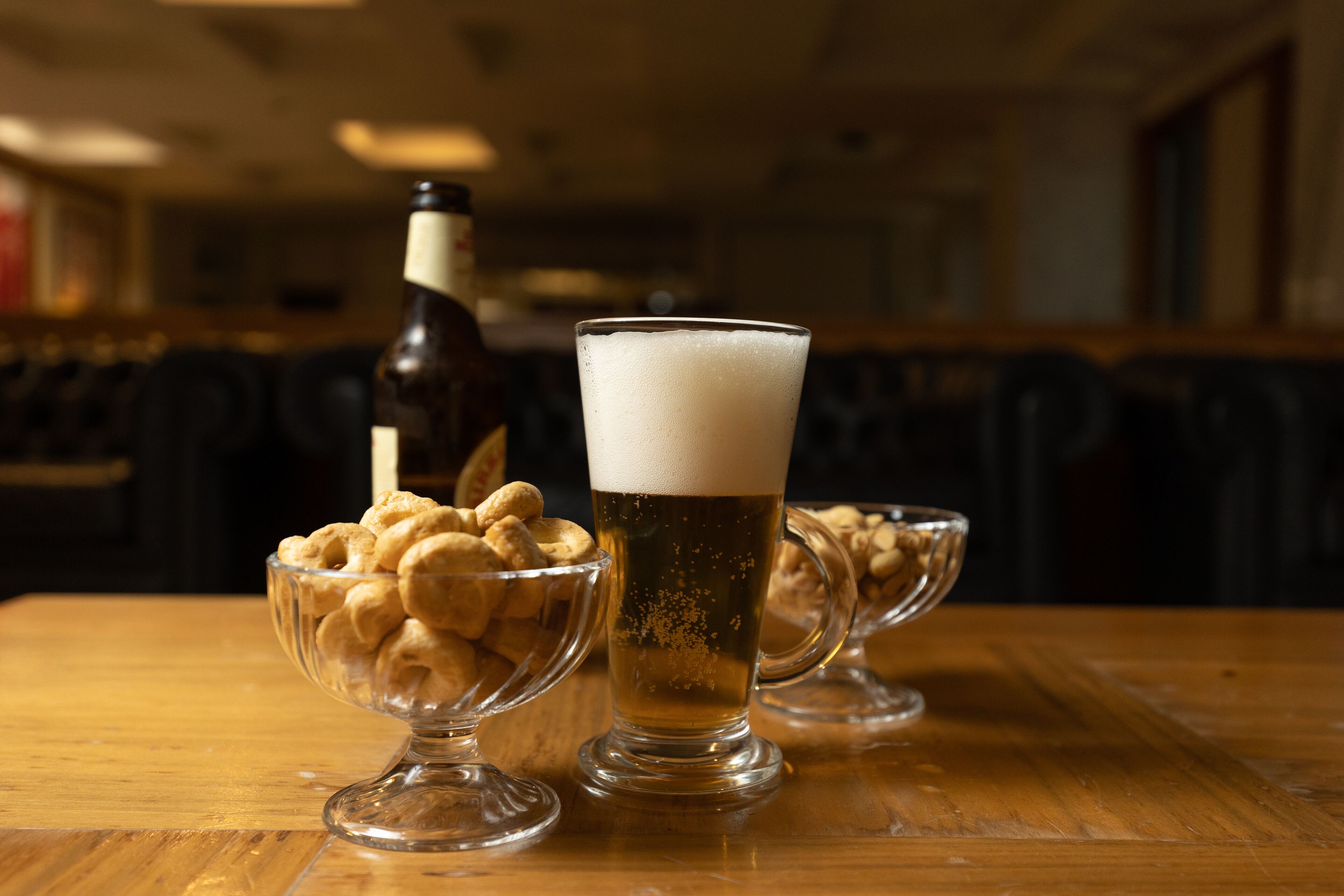 A glass of beer on a wooden table with two bowls of peanuts.