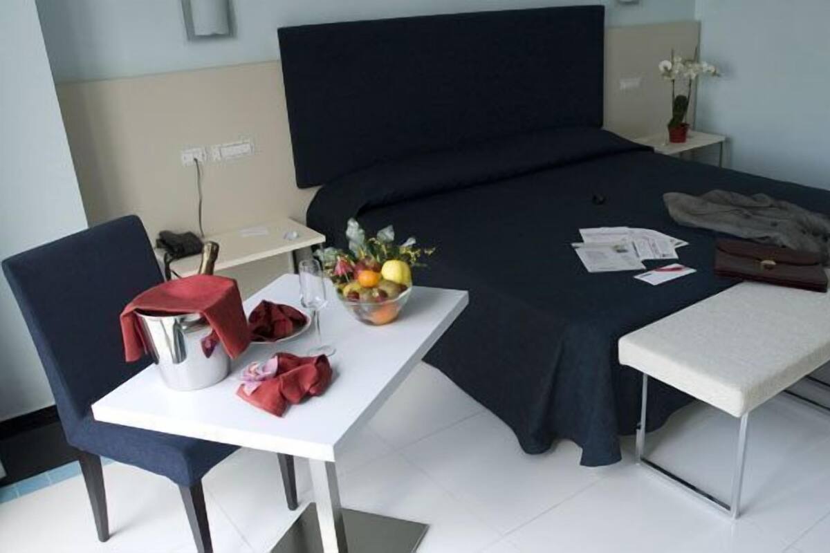Habitación de hotel con cama, una mesita con frutero, una silla, un banco y una mesita de noche con lámpara.
