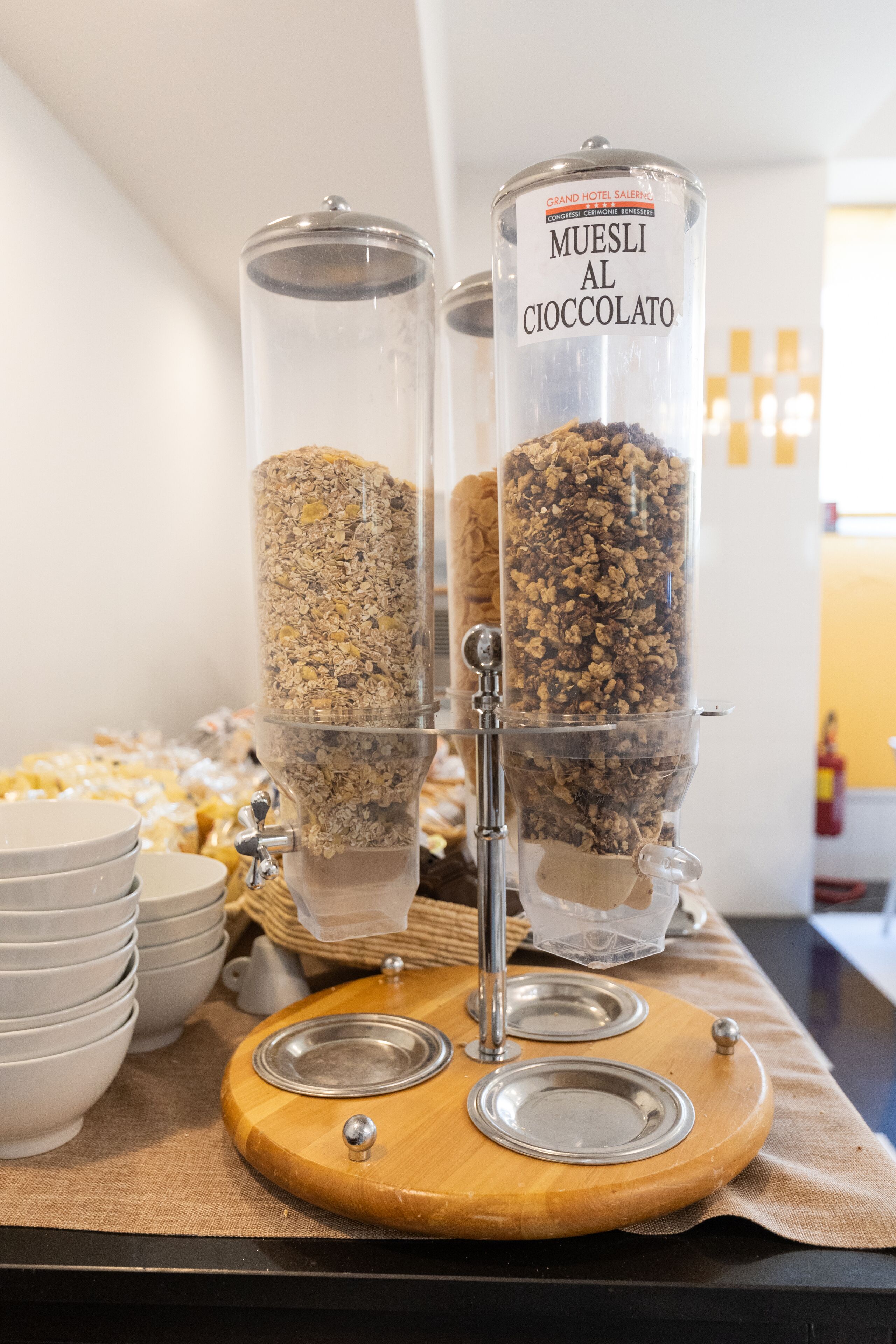 Två behållare märkta med texten ”MUESLI AL CIOCCOLATO” står på en träbräda, och under dem finns skålar och tallrikar.