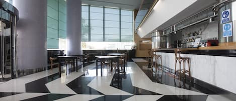 Un moderno vestíbulo de hotel con suelo a cuadros blanco y negro, amplias ventanas y una zona de bar con taburetes.