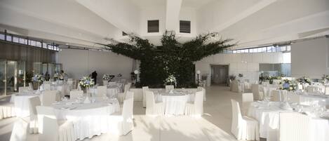 Un salón amplio con varias mesas redondas dispuestas para un evento formal, con manteles blancos y sillas, y una planta decorativa grande en el centro.