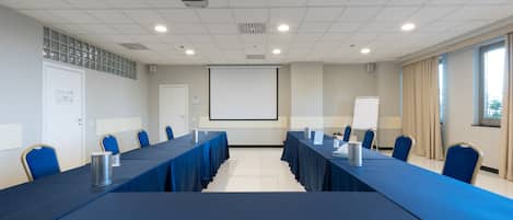 Un salón de conferencias con una larga mesa, sillas azules, una pantalla de proyector y una pizarra.