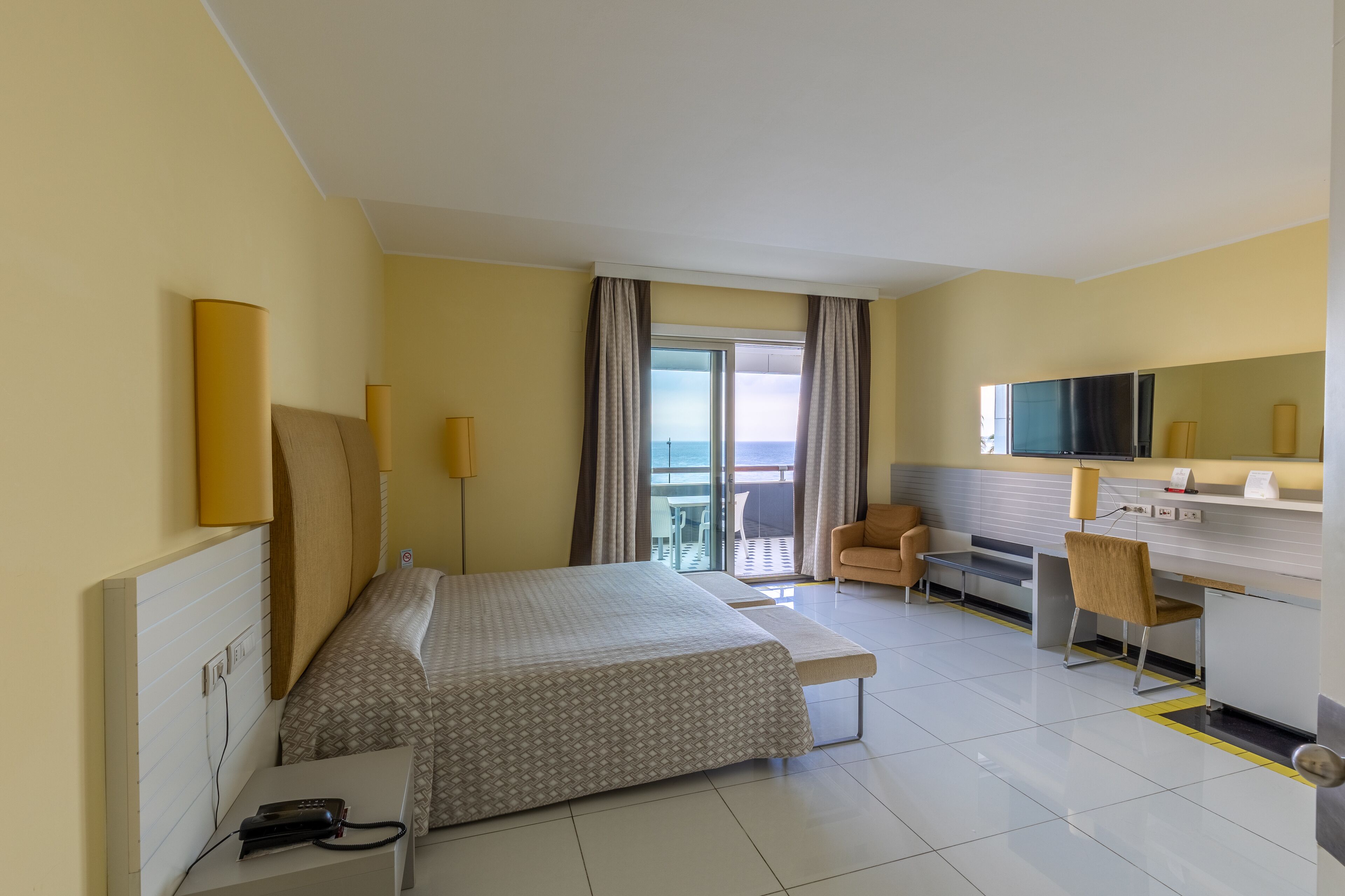 A hotel room with a bed, desk, chair, and a view of the sea.