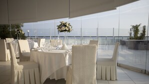 Ein Essbereich mit einem für sechs Personen gedeckten Tisch, umgeben von weißen Stühlen, mit Blick auf das Meer durch große Fenster.