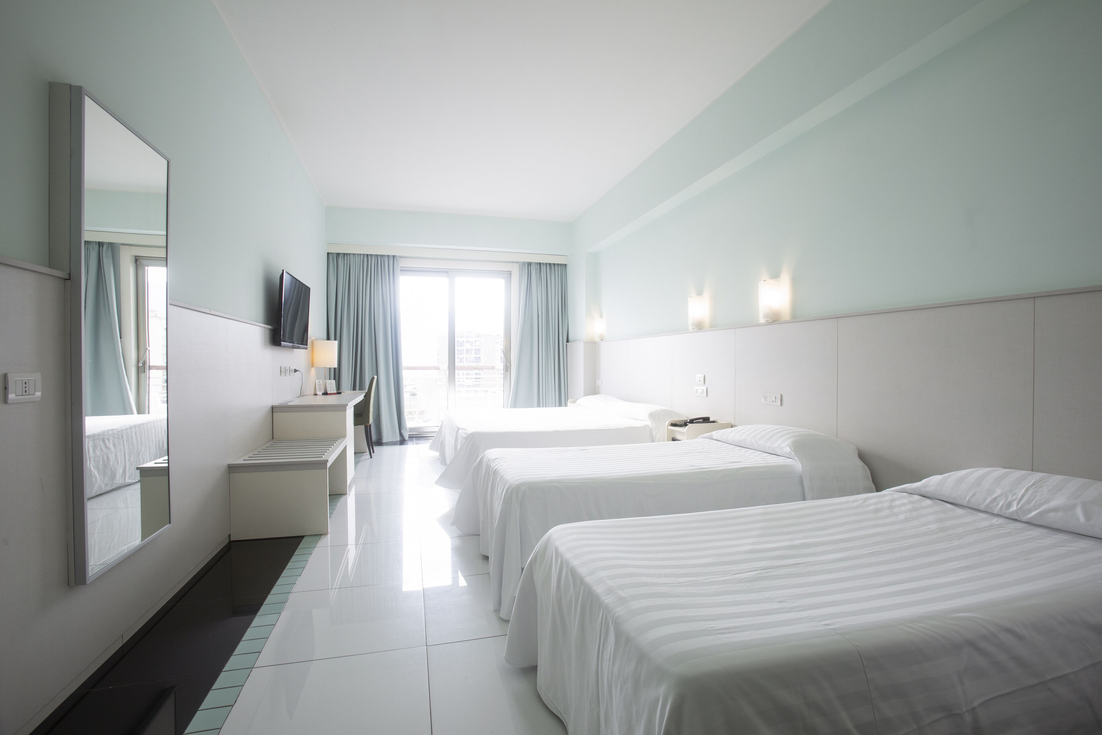 Ein Hotelzimmer mit drei Betten, einem Fernseher, einem Schreibtisch und einem Fenster mit Vorhängen.