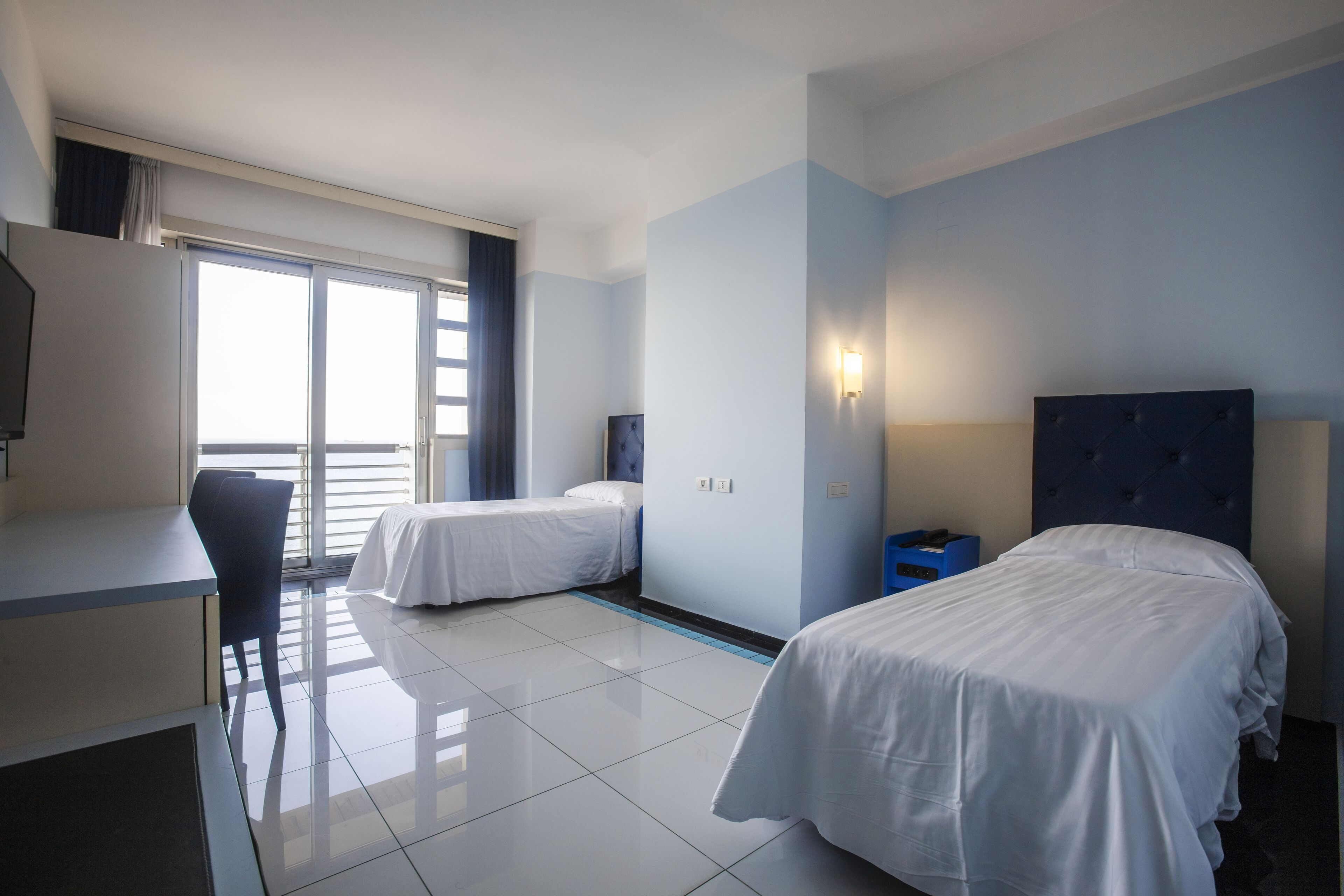 Una camera d'albergo con due letti, una scrivania, una sedia e un'ampia finestra.