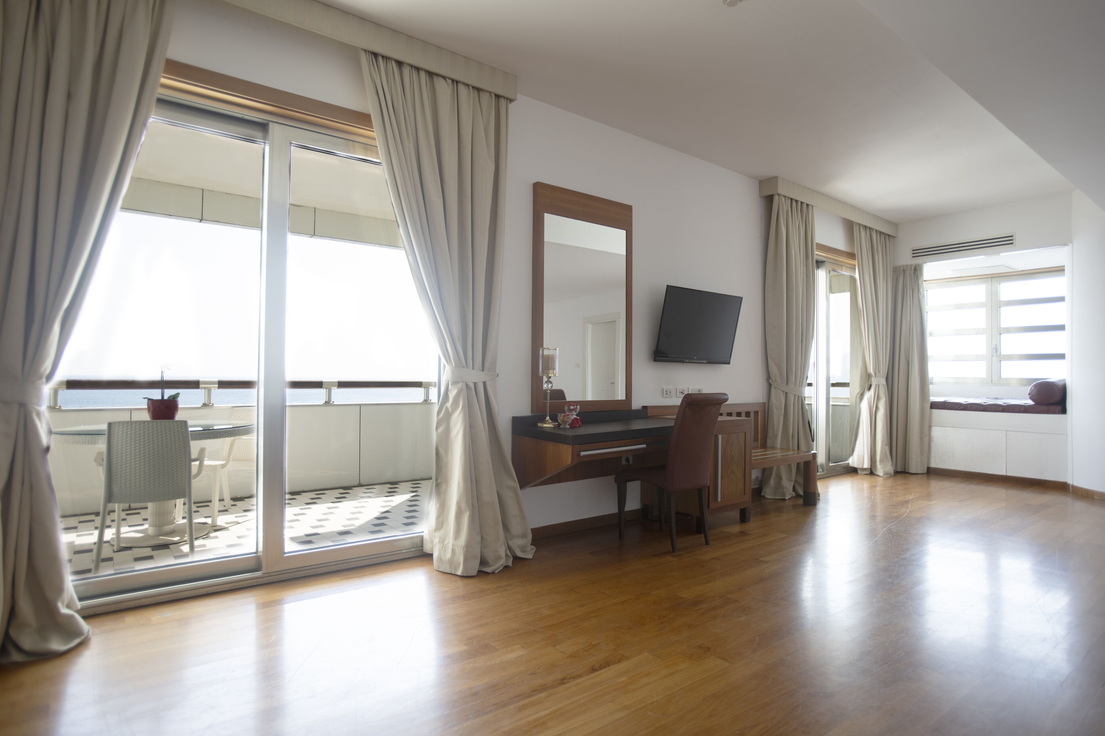 Una habitación amplia con un gran ventanal corredero que da a un balcón, un escritorio de madera con una silla, un televisor montado en la pared y una ventana con cortinas.