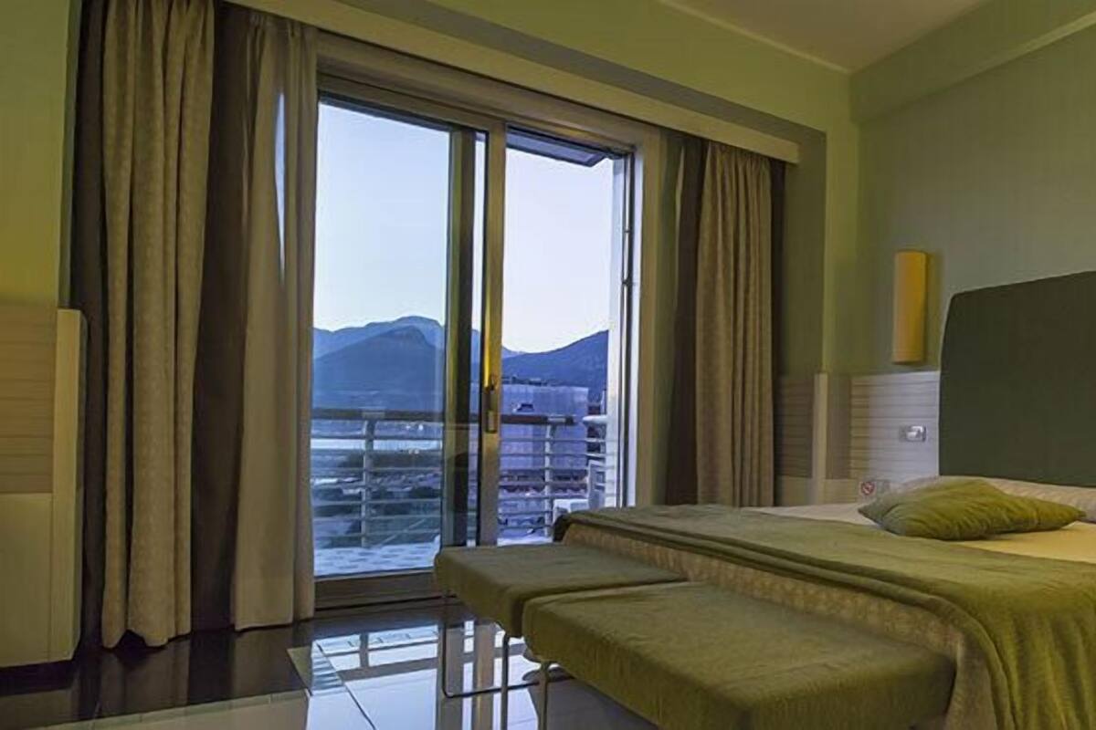 Habitación de hotel con una cama grande, un banco, una mesita de noche y vistas a montañas a través de un ventanal.