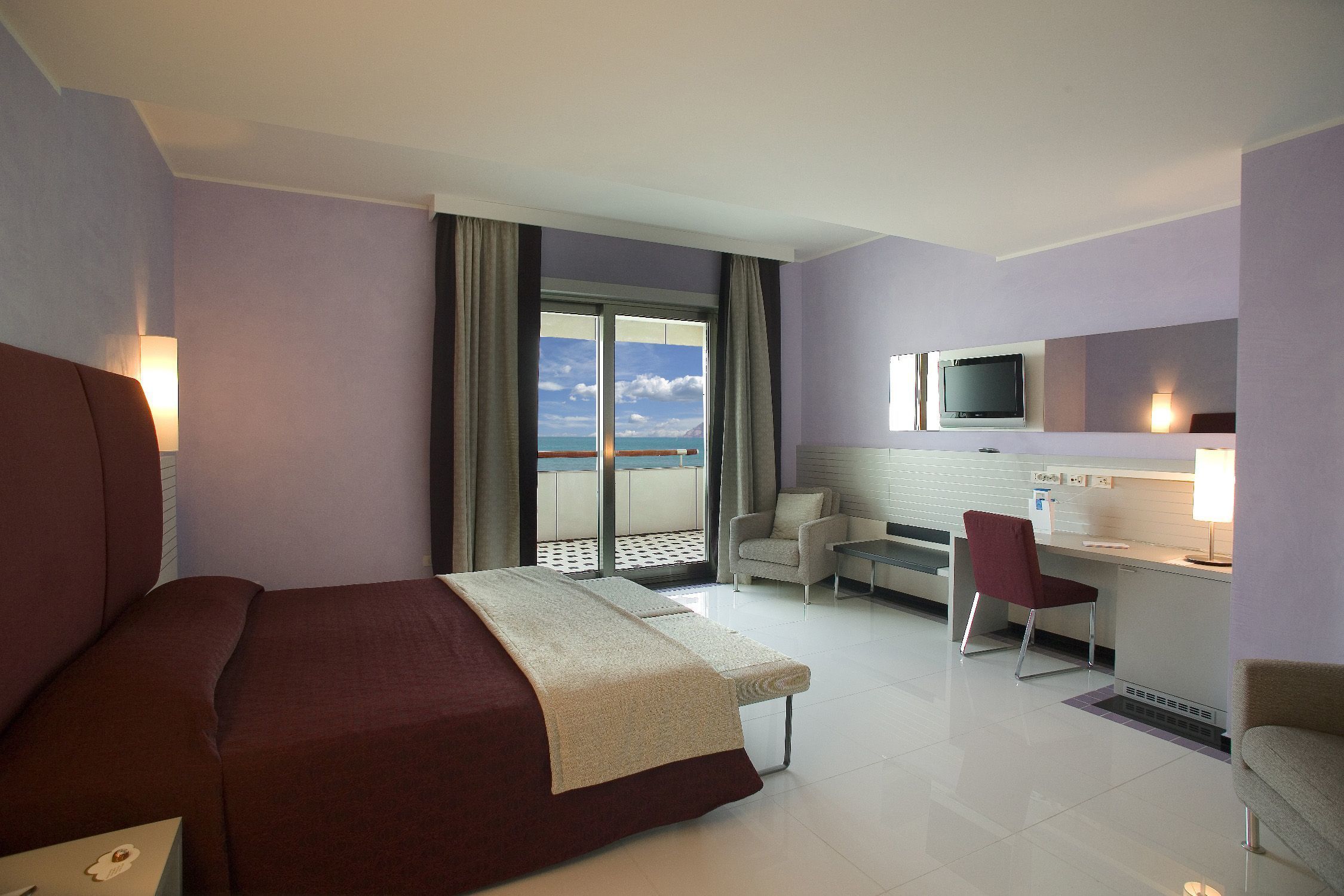 Habitación de hotel con cama, escritorio, silla, televisor y vista al mar.
