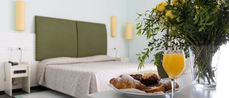 Hotelkamer met een bed, nachtkastje en een ontbijtarrangement, waaronder croissants, een glas sinaasappelsap en een bloevas.