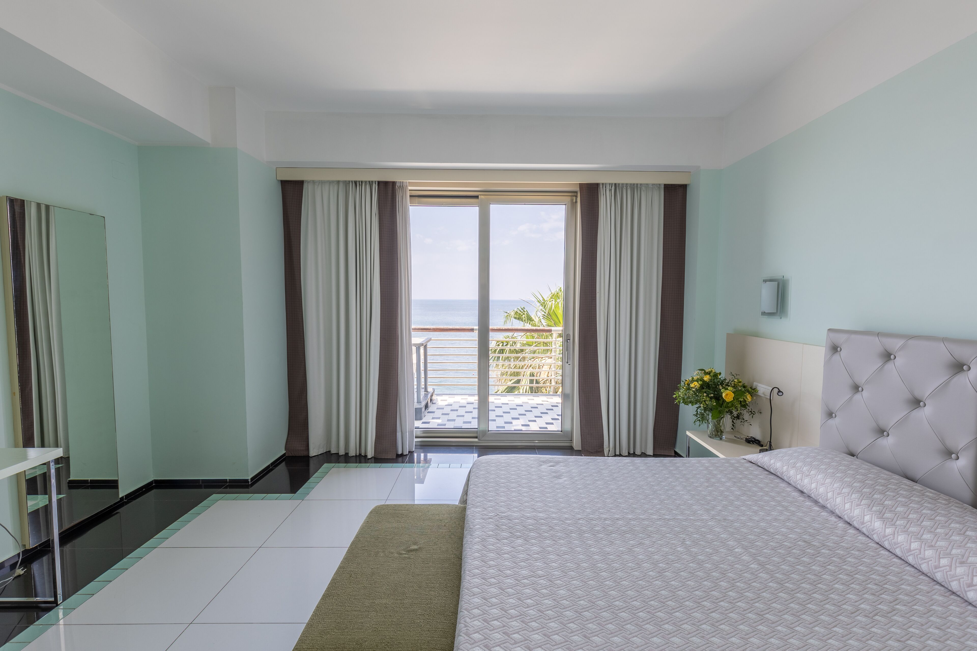 Une chambre d’hôtel avec un lit, une table de chevet, un miroir et une vue sur l’océan, accessible par une porte coulissante.