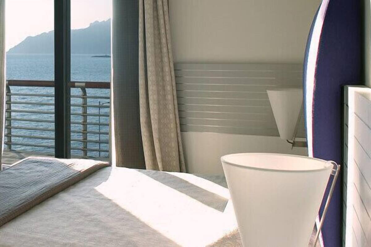 A bedroom with a large window, a bed, a chair, and a bedside table.
