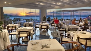 Een restaurant met gedekte tafels, grote ramen met uitzicht op de zonsondergang en de zee, en een modern interieur.