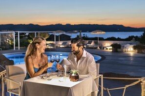 Un couple est assis à une table près d’une piscine, avec vue sur les montagnes et un plan d’eau, au crépuscule.