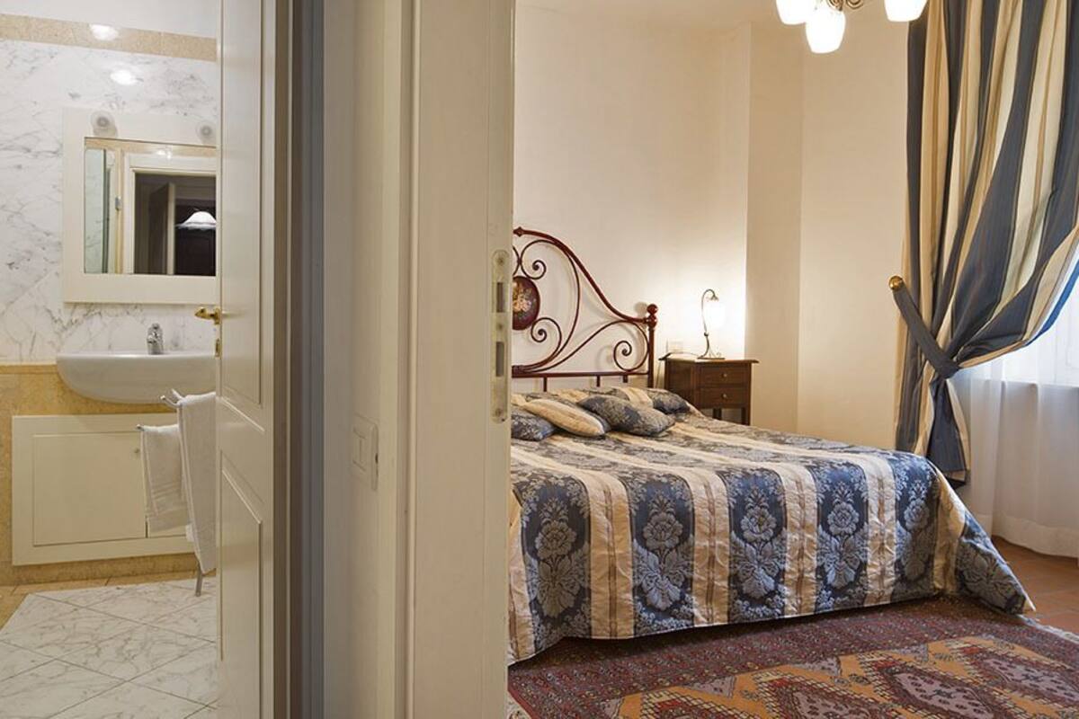 Een slaapkamer met een bed, nachtkastje, spiegel, raam met gordijnen en een badkamer met wastafel en toilet.