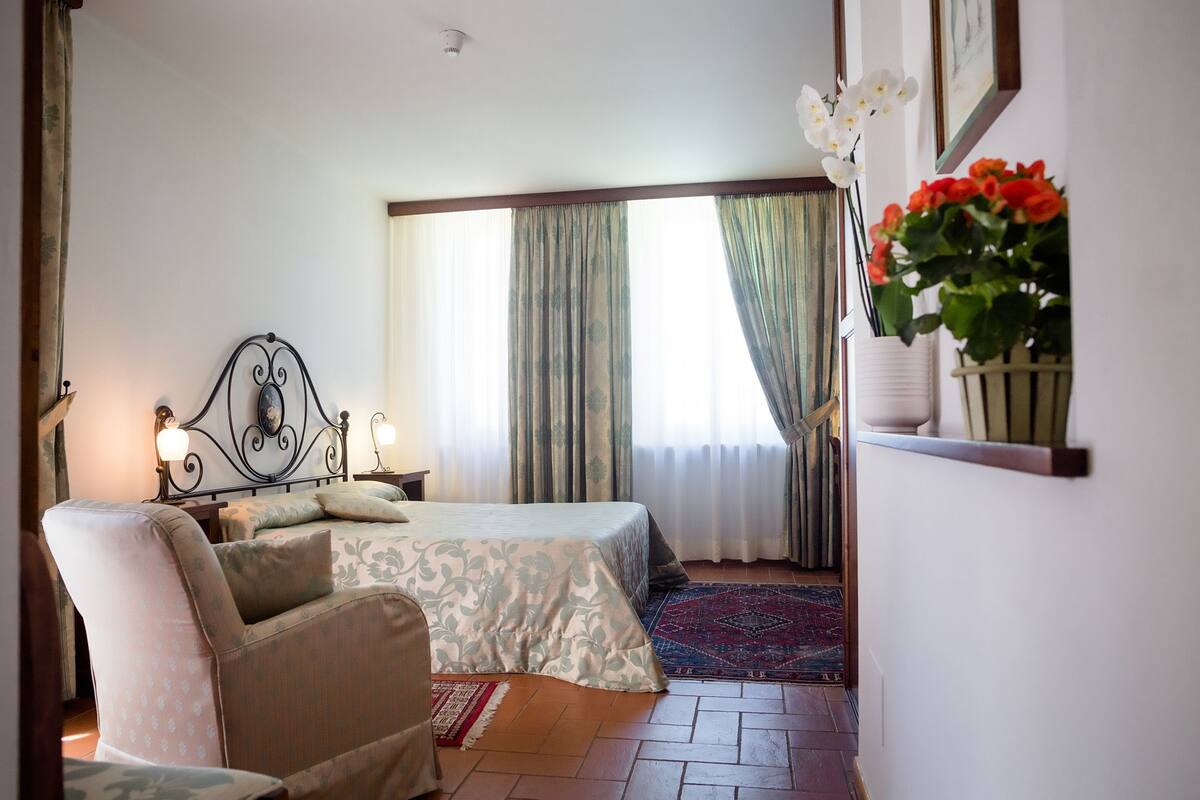 Een slaapkamer met een bed, een stoel, een nachtkastje, een raam met gordijnen en een bloempot met plant.