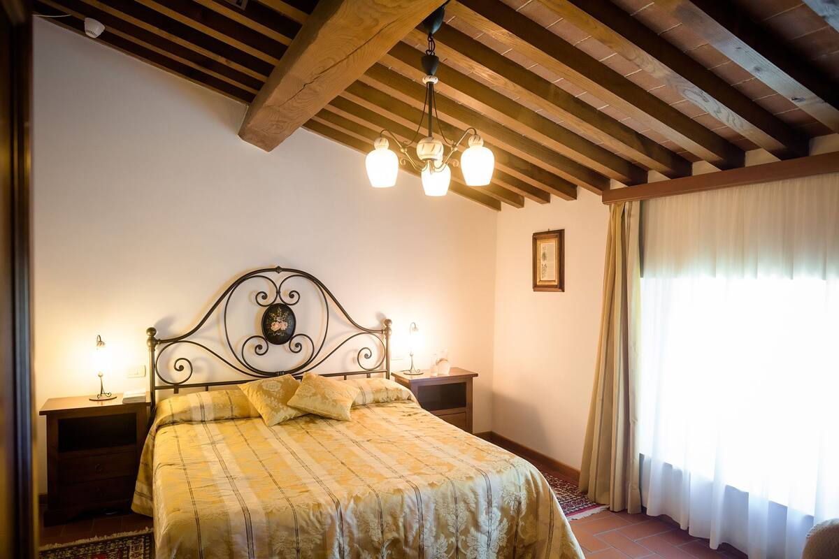 Een slaapkamer met een bed, houten balken in het plafond, een kroonluchter, een nachtkastje en een raam met gordijnen.