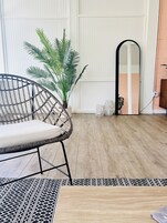 A room with a wooden floor, a black woven chair, a large potted plant, a mirror, and a patterned rug.
